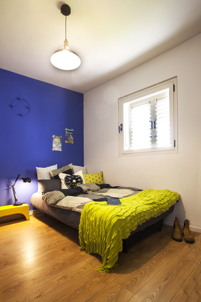 חדר ממ״ד, עם קיר צבוע כחול רואייל, מיטה וחצי עם כריות בגוונים שחור לבן ואפור כהה. 