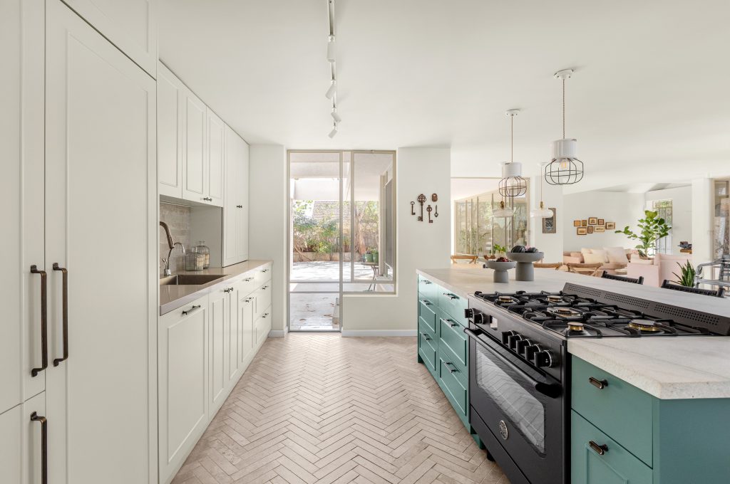 המטבח של קוקו שאנל. מטבח בגוון בהיר עם אי בגוון ירוק ים, כיסאות בר שחורים בשילוב חבל שחור. חיבור מעוגל בין פרקט מעושן כהה לאריח הרינגבון בגוון חול טבעי.