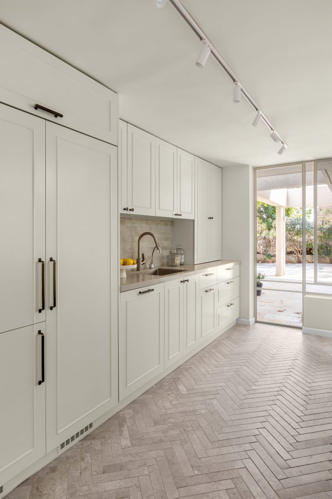 מטבח בגוון בהיר ארונות גבוהים וכיור. מקרר אינטגרלי, אריח אבן טבעי הרינגבון בגוון חול, חלונות בלגיים.