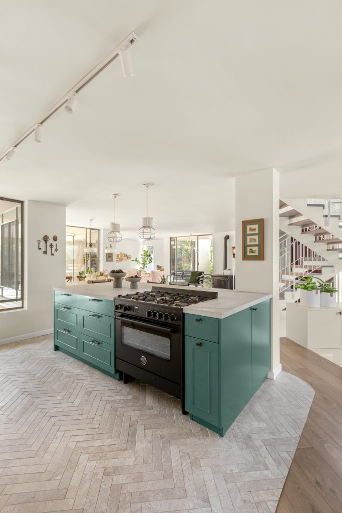 מטבח, מבט אל אי בגוון ירוק ים, כיסאות בר שחורים בשילוב חבל שחור. חיבור מעוגל בין פרקט מעושן כהה לאריח הרינגבון בגוון חול טבעי. מנורות אמייל תעשייתיות. תנור עומד שחור.